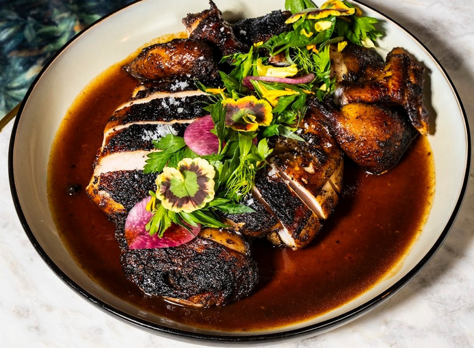 A plate of sliced, pimento-smoked jerk chicken, topped with herbs and sitting in a dark red jus.