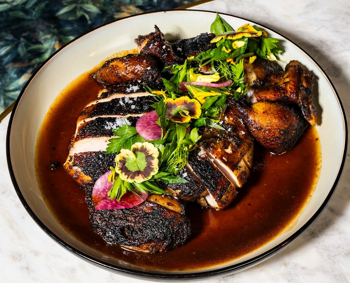 A plate of sliced, pimento-smoked jerk chicken, topped with herbs and sitting in a dark red jus.