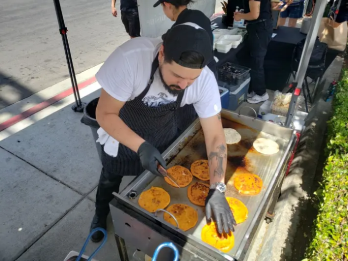 A chef in a backwards baseball cap managing ten pupusas on a hot griddle