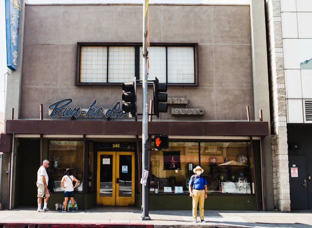 an image of a sidewalk and building, the Bunkado shop. three people are outside.