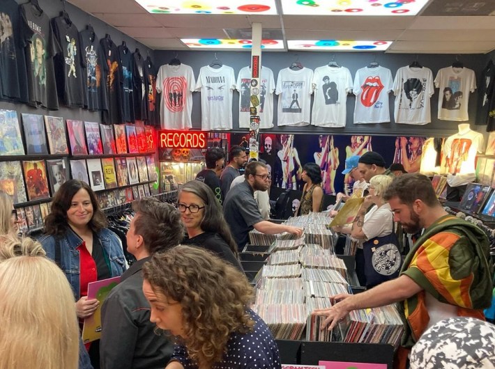 in a record shop, many people are crowded as the browse records or mingle. there are band and artist t-shirts hanging on the walls.