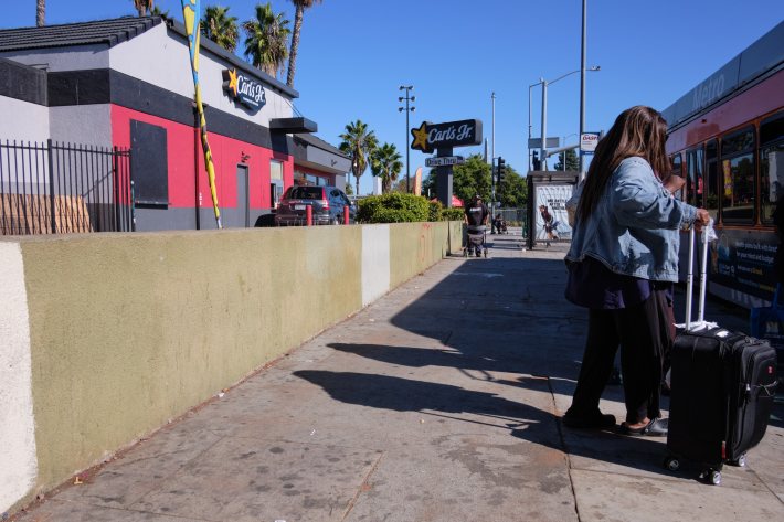 sidewalk between a bus and carl's jr. a woman is holding onto a suitcase