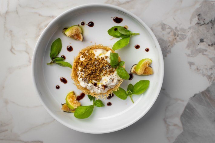 A white plate with an artistic dish of burratina in a crown of pastry with crumbled almonds on top, surrounded by leaves of basil and dots of balsamic