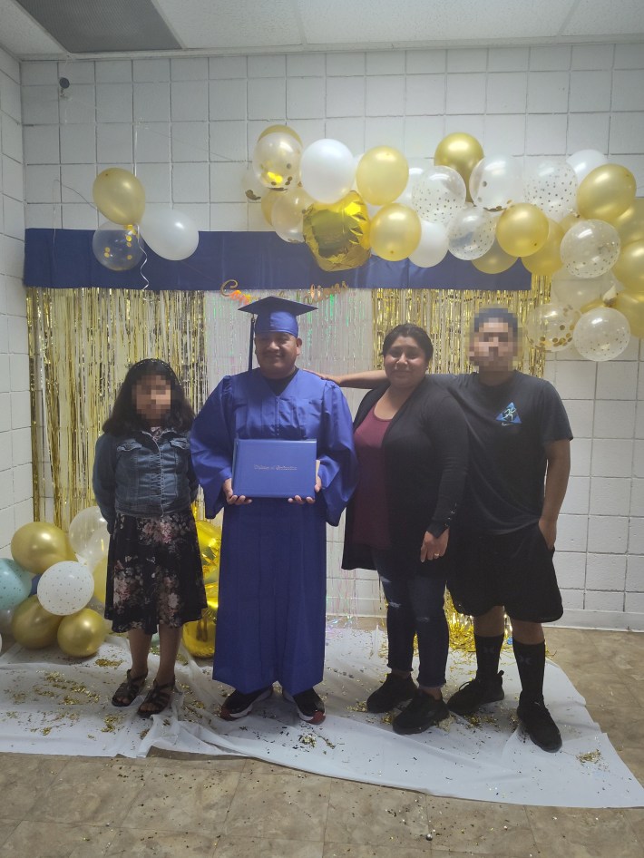A young man in a graduation gown stands with two siblings and his mother in front of gold tinsel curtains and balloons.