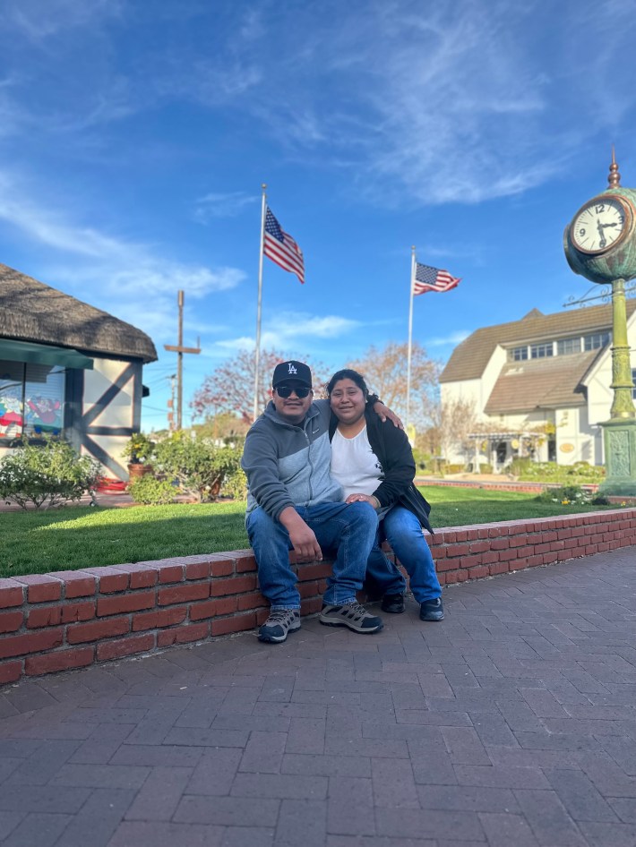 A man in a Dodgers hat has his arm around a woman, both smike, as two U.S. flags flutter above them