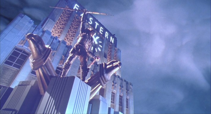 an alien stands on a roof platform raising his weapon in the air