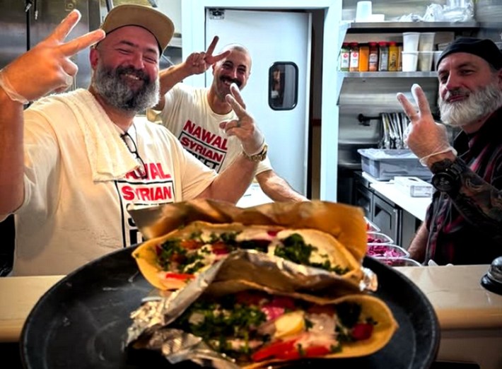 Three men flashing peace signs, with a blurry serving of falafels in the foreground, on a counter.