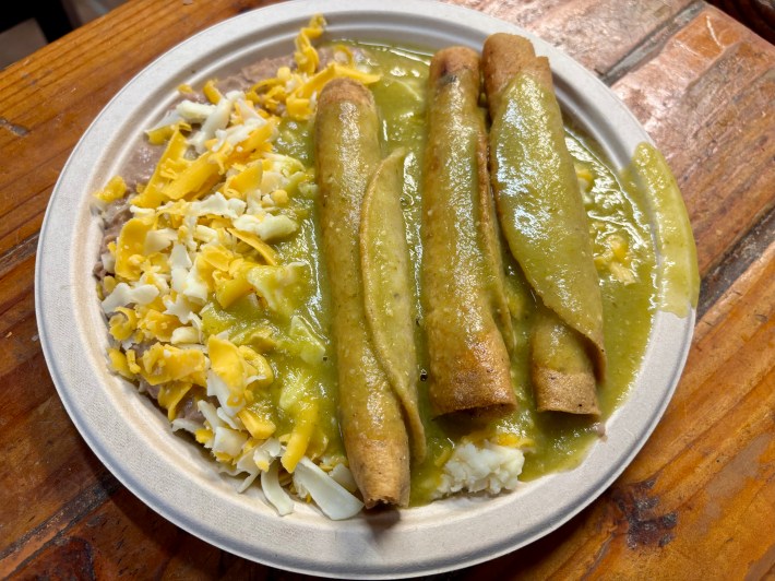 a plate of taquitos in green sauce with beans and cheese on the side