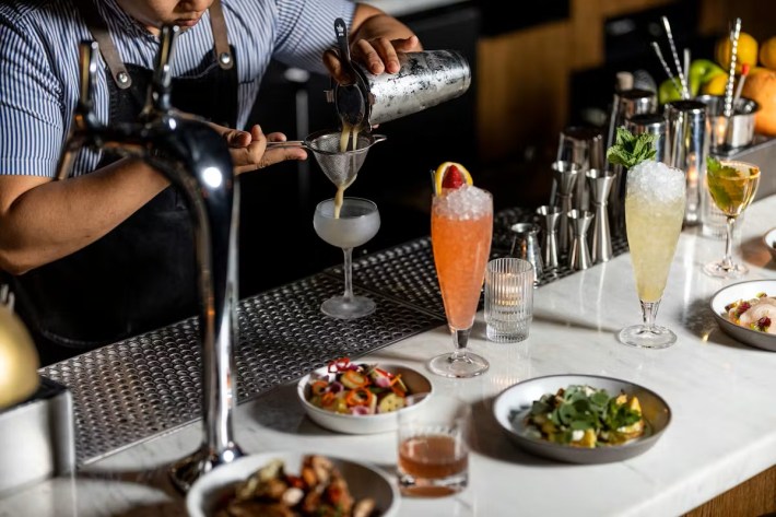 A bartender strains a cocktail, while other cocktails and plates of food sit before him on a marble bar