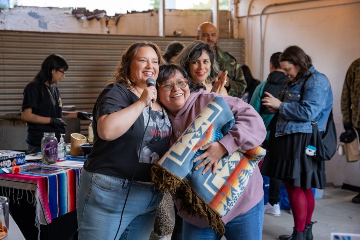 two women hug and smile as another one behind them claps. one woman is holding a microphone