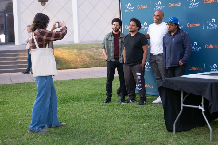 A man poses for a photographer beside three younger men on a lawn, with a CalTech backdrop behind them.