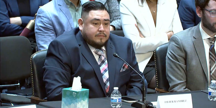 a man sits in a suit at a desk, a box of tissues near him