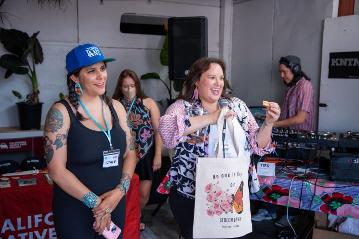 two women smiling. one is wearing a blue cap. the other is holding a tote bag. a dj plays a set behind them