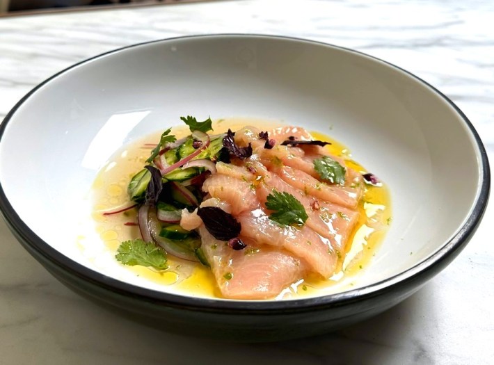 A plate of yellowtail crudo in a pool of yellow oil and toipped with herbs