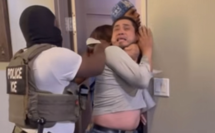 an ICE agent wearing a tshirt, vest, and head covering holds onto a person who is smushed against a man with a look of terror on his face