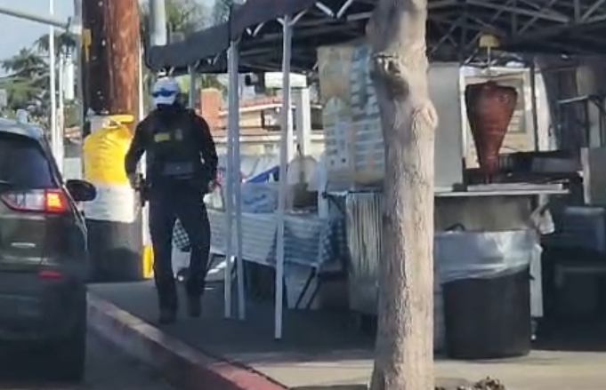 an armed border patrol agent lurks near a food stand on the sidewalk
