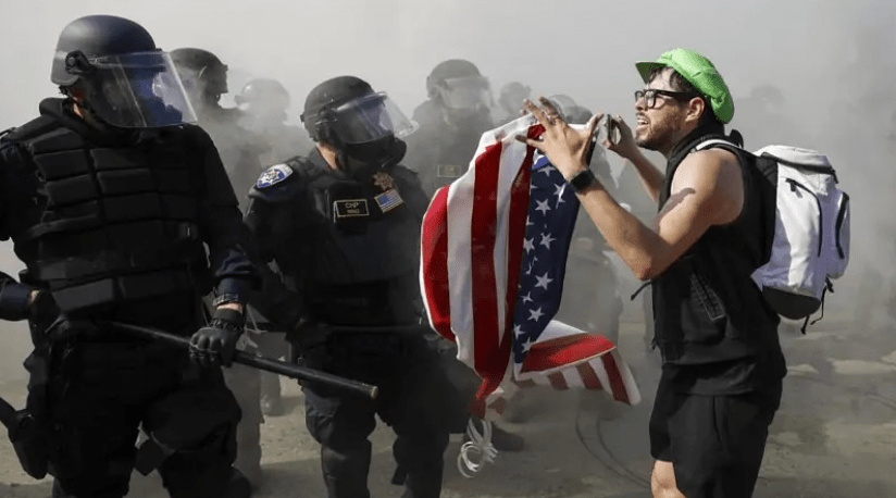a man holds an American flag while protesting in front of armed officers in riot gear