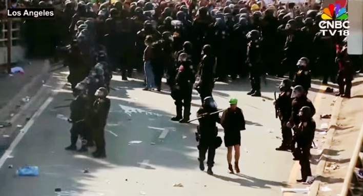 a man in a green Luigi hat is arrested. a large crowd of officers armed and wearing riot gear are behind him.