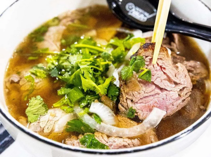 A bowl of pho with chopsticks lifting a cut of steak, with onions and cilantro