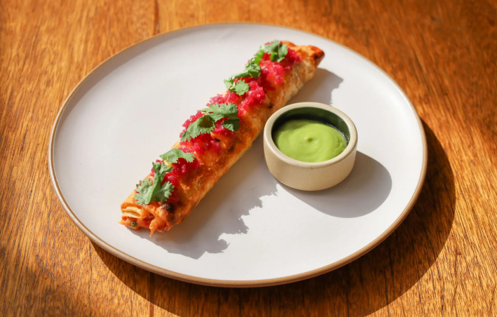 A red-sauced flauta with smoked tuna and queso Chihuahua and a bowl of green sauce on a white plate