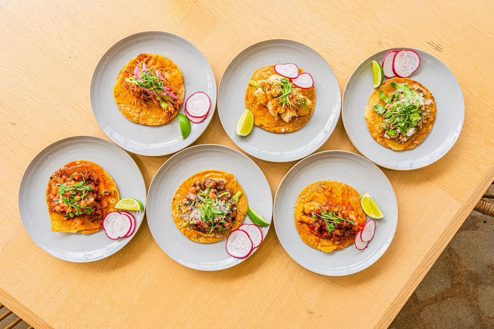 Six white plates, each holding a taco, on a wooden table