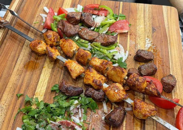 Skewered beef and chicken kebab on a wooden cutting board next to tomatoes and herbs