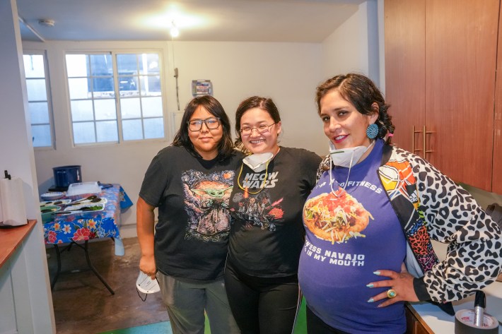 three women stand together, posing while wearing safety masks. one woman is pregnant
