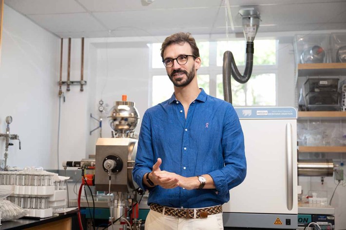 A man in glasses and a blue shirt speaks in front of a science lab