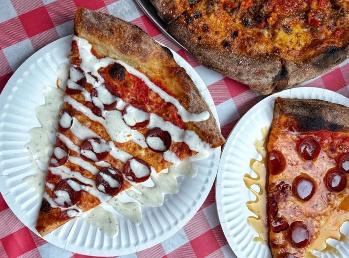 Two slices of pepperoni pizza on paper plates, one with white sauce drizzled all over it, and a part of a pizza pie peeking in from the top