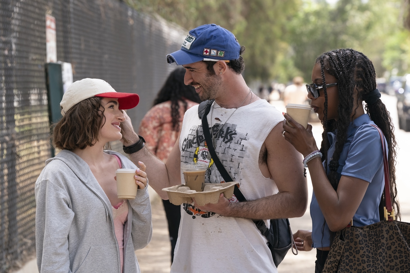 Three stars from I Love LA talking in front of a fence while holding coffee, in a still from the show