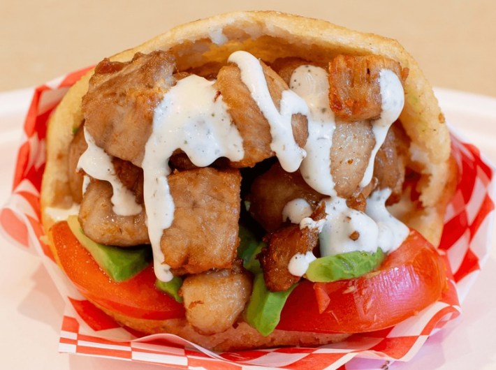 A paper-wrapped sandwich bursting with pork belly, white sauce, and tomato
