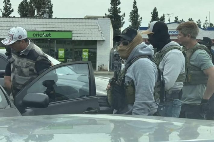 Masked and unmasked men wearing tactical gear near a car in a plaza parking lot