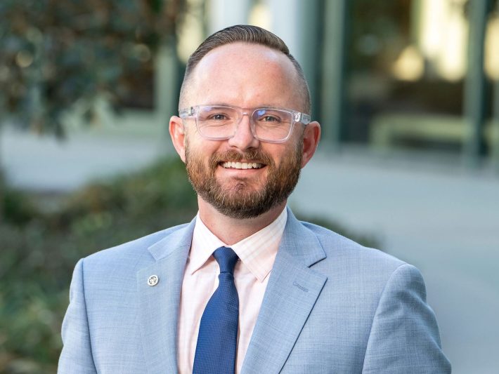 a man with glasses and a beard wearing a suit smiles at the camera