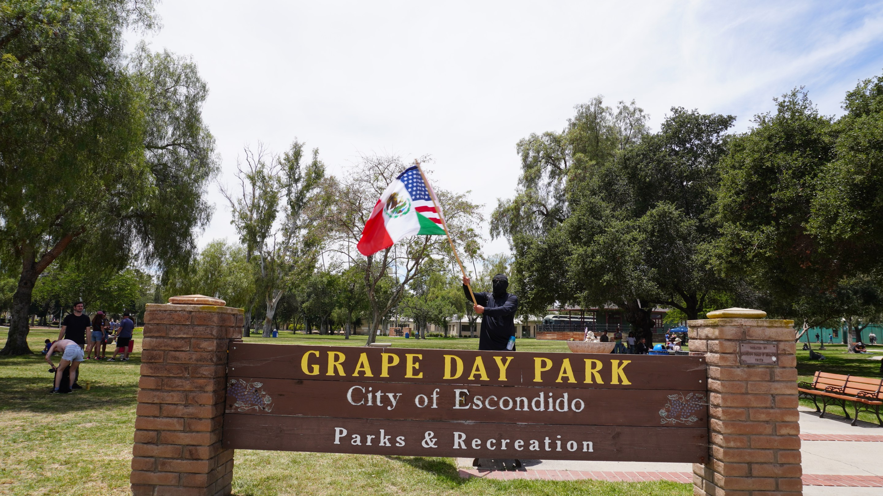 a person waves a flag (which is a mashup of the Mexica and American flag) in front of a sign that says "Grape Day Park City of Escondido"