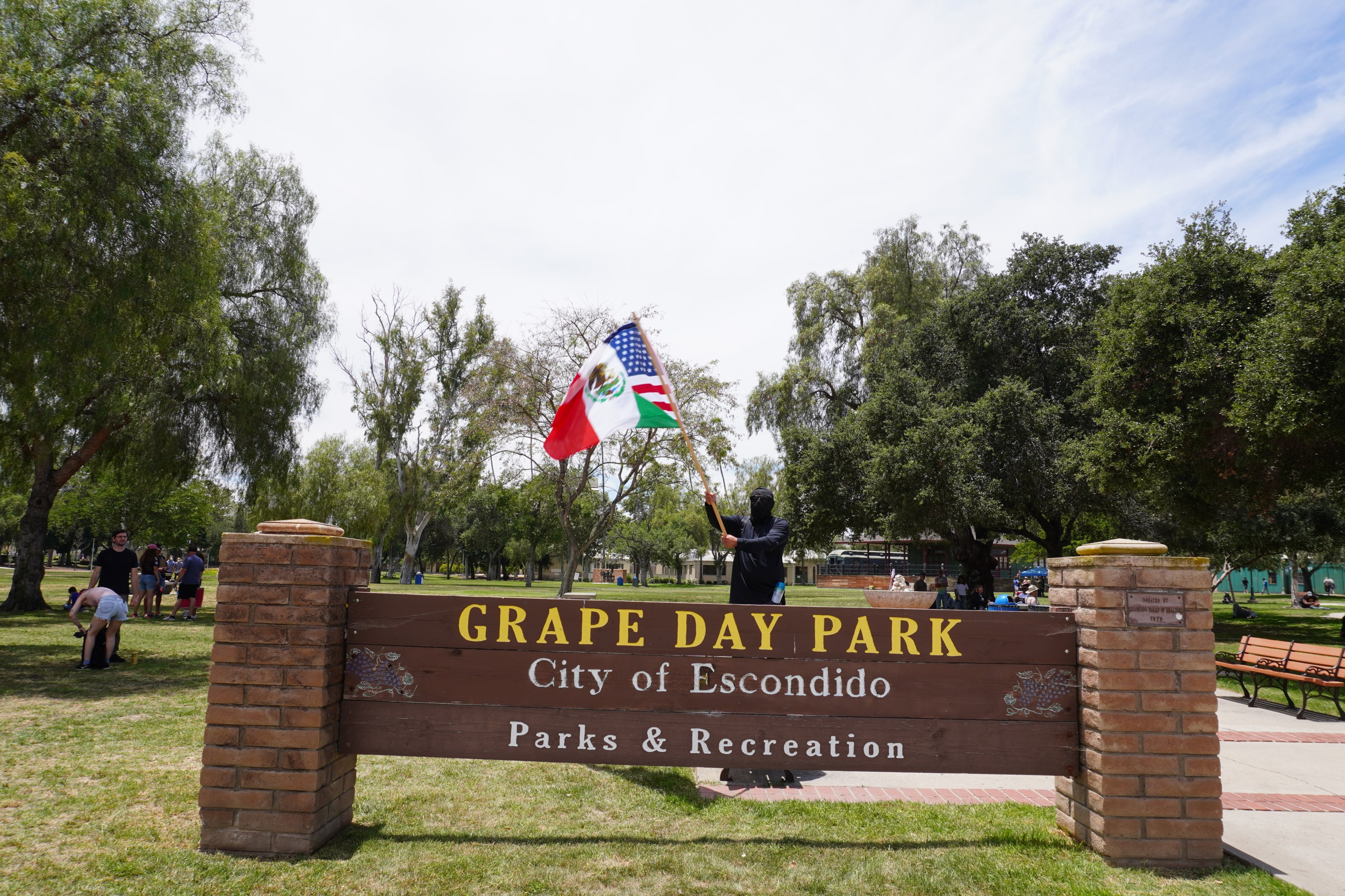 a person waves a flag (which is a mashup of the Mexica and American flag) in front of a sign that says "Grape Day Park City of Escondido"