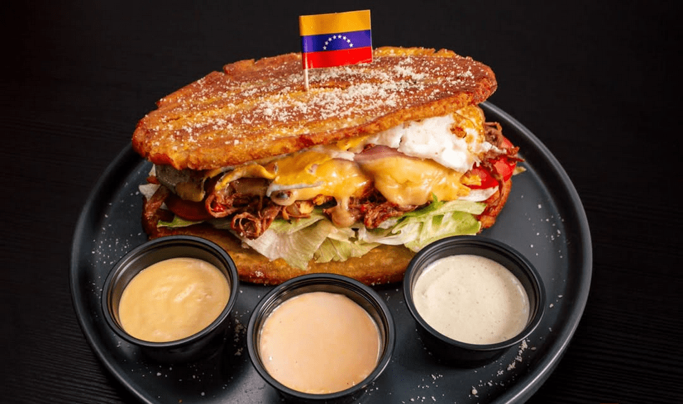 A massive sandwich with a tiny Venezuelan flag on top, ringed by 3 sauces