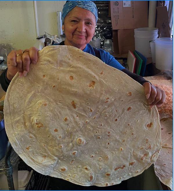 A woman in a kerchief holds a massive flour tortilla she's made.