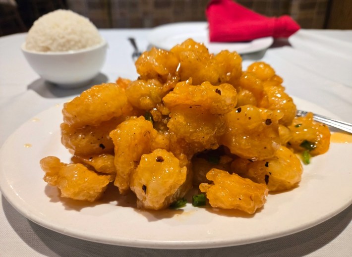 A white plate of orange slippery shrimp, with a bowl of rice and a red napkin behind it