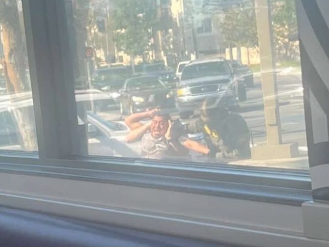 a photo taken through a window of a home. outside, a man is being forcefully held by a federal agent