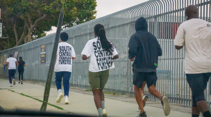 six people running on a sidewalk, two wearing shirts that read "SOUTH CENTRAL FOREVER" on the back