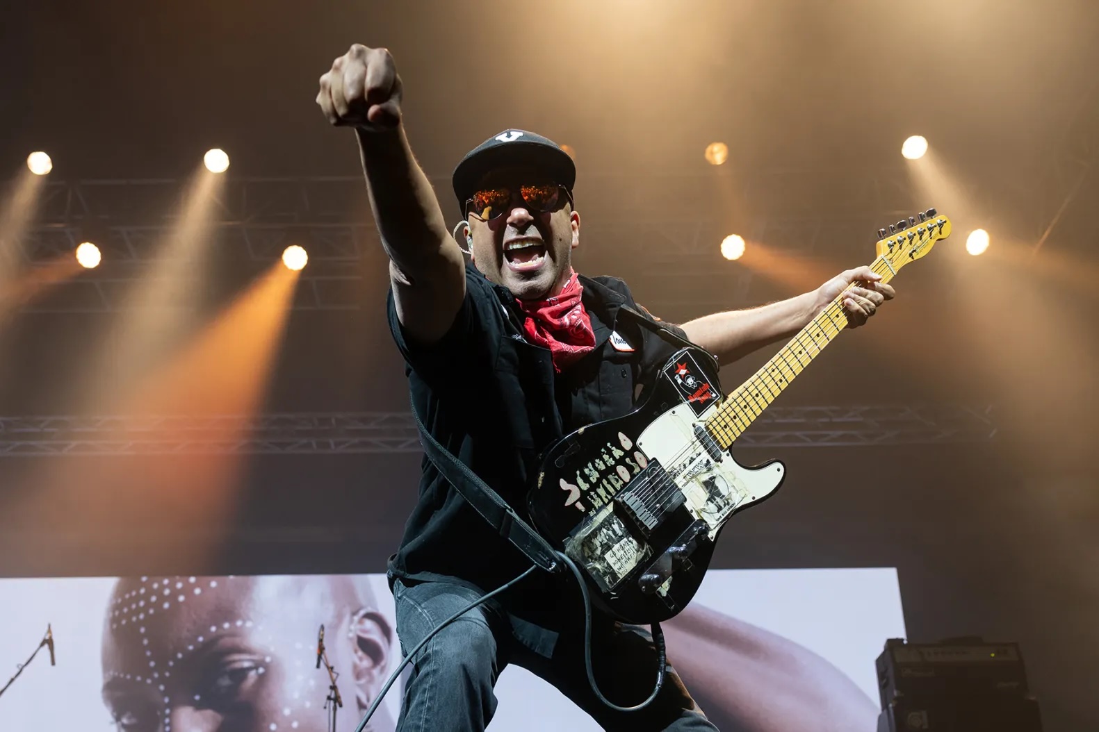 a man onstage wears a hat and holds a guitar while raising a fist in the air and smiling