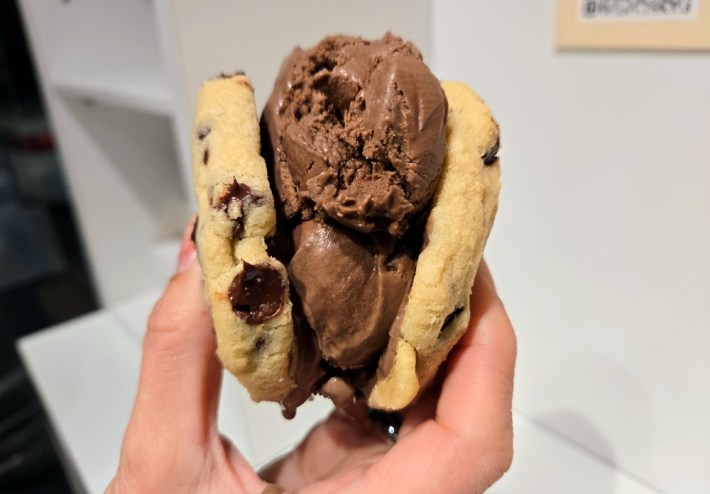 An ice cream sandwich with two chocolate chip cookies sandwiching chocolate ice cream, held in someone's hand.
