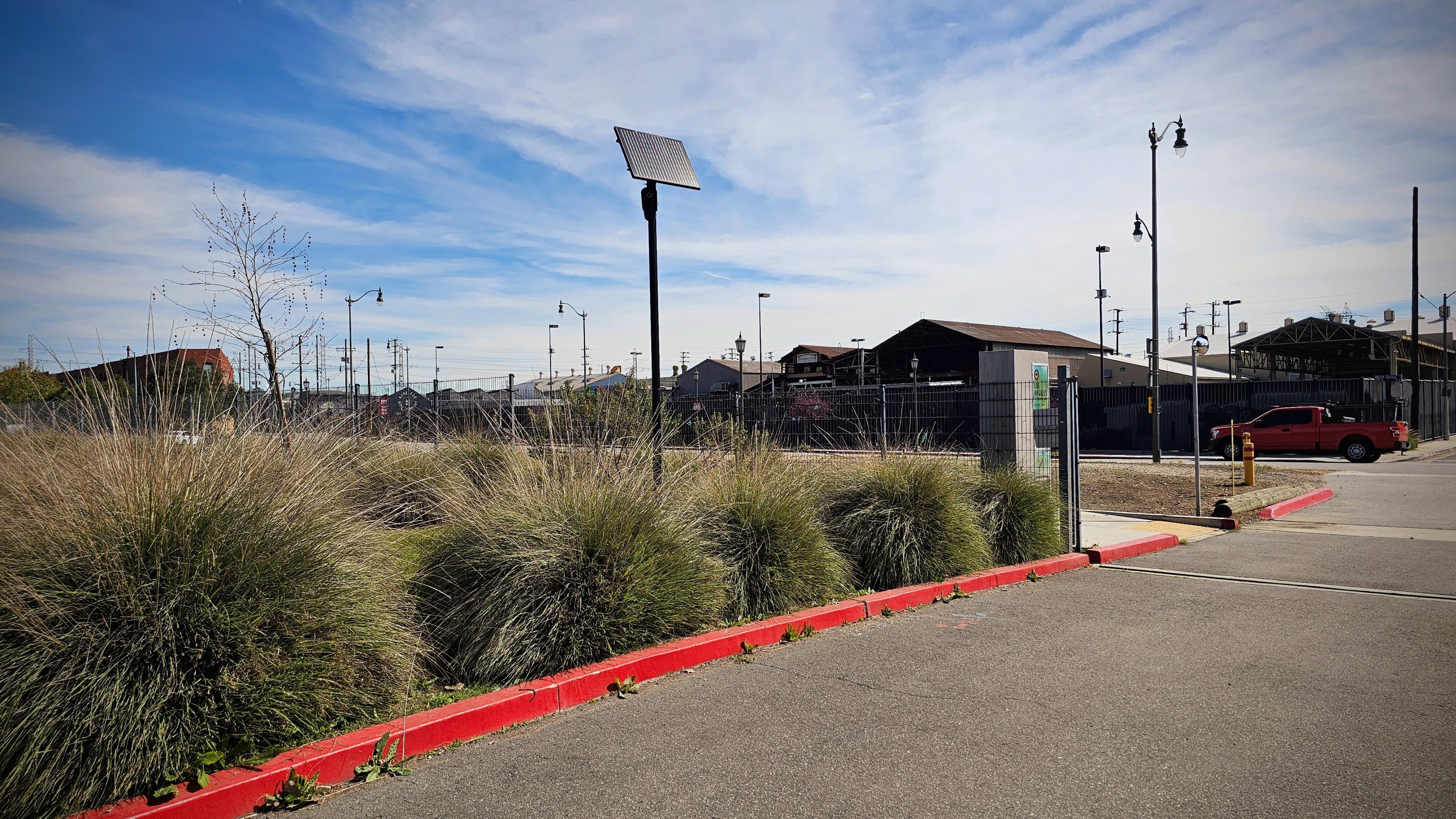 A Flock license plate reader behind a row of shrubs near the entrance of L.A. State Historic Park. A car drives past the park in the background.