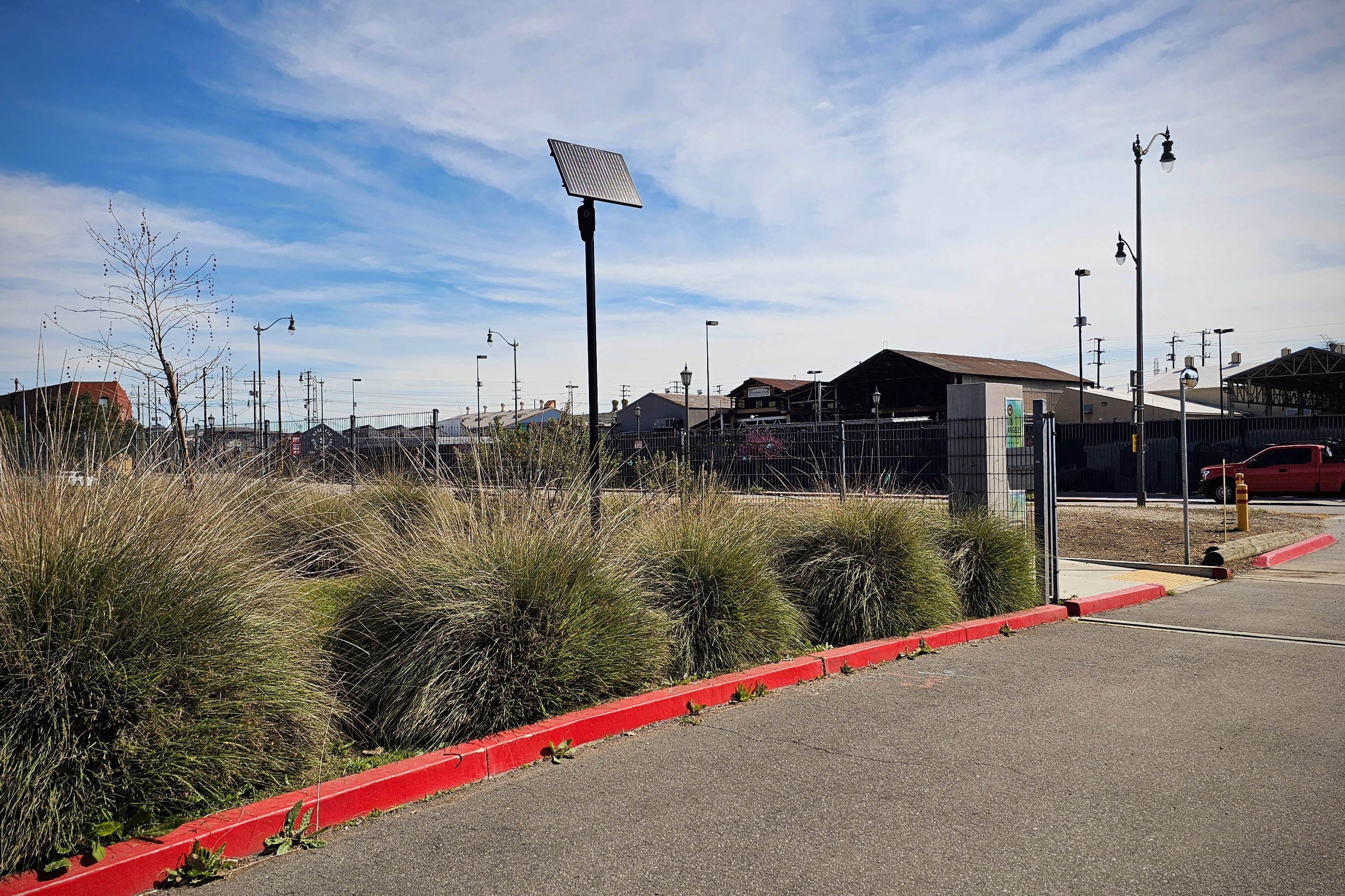 A Flock license plate reader behind a row of shrubs near the entrance of L.A. State Historic Park. A car drives past the park in the background.