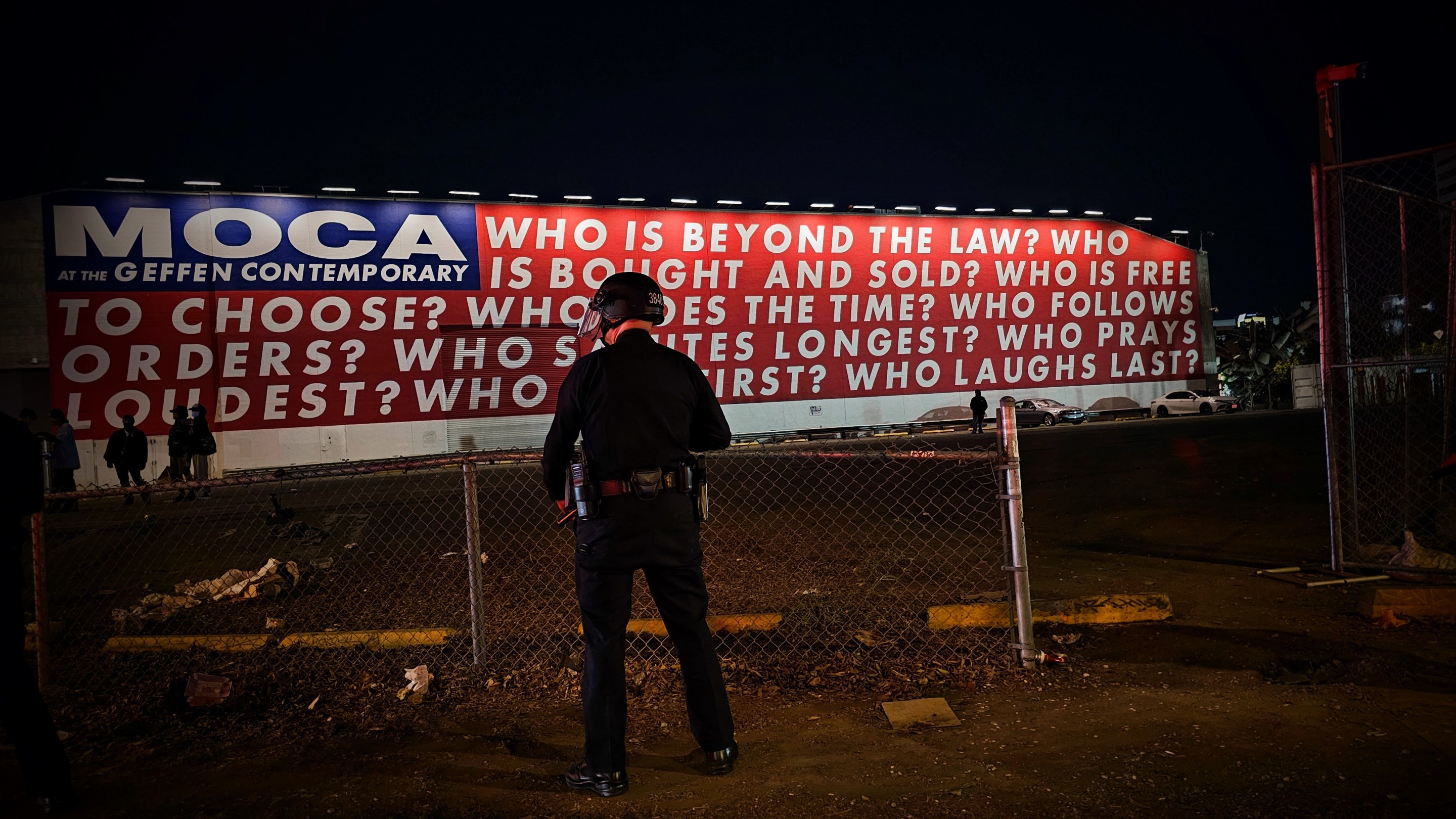 A Los Angeles police officer stands facing a red, white, and blue mural by the artist Barbara Kruger. The mural asks nine questions: “Who is beyond the law?”, “Who is bought and sold?”, “Who is free to choose?” “Who does the time?”, “Who follows orders?”, “Who Salutes longest?”, “Who prays loudest?”, “Who dies first?” and “Who laughs last?”.