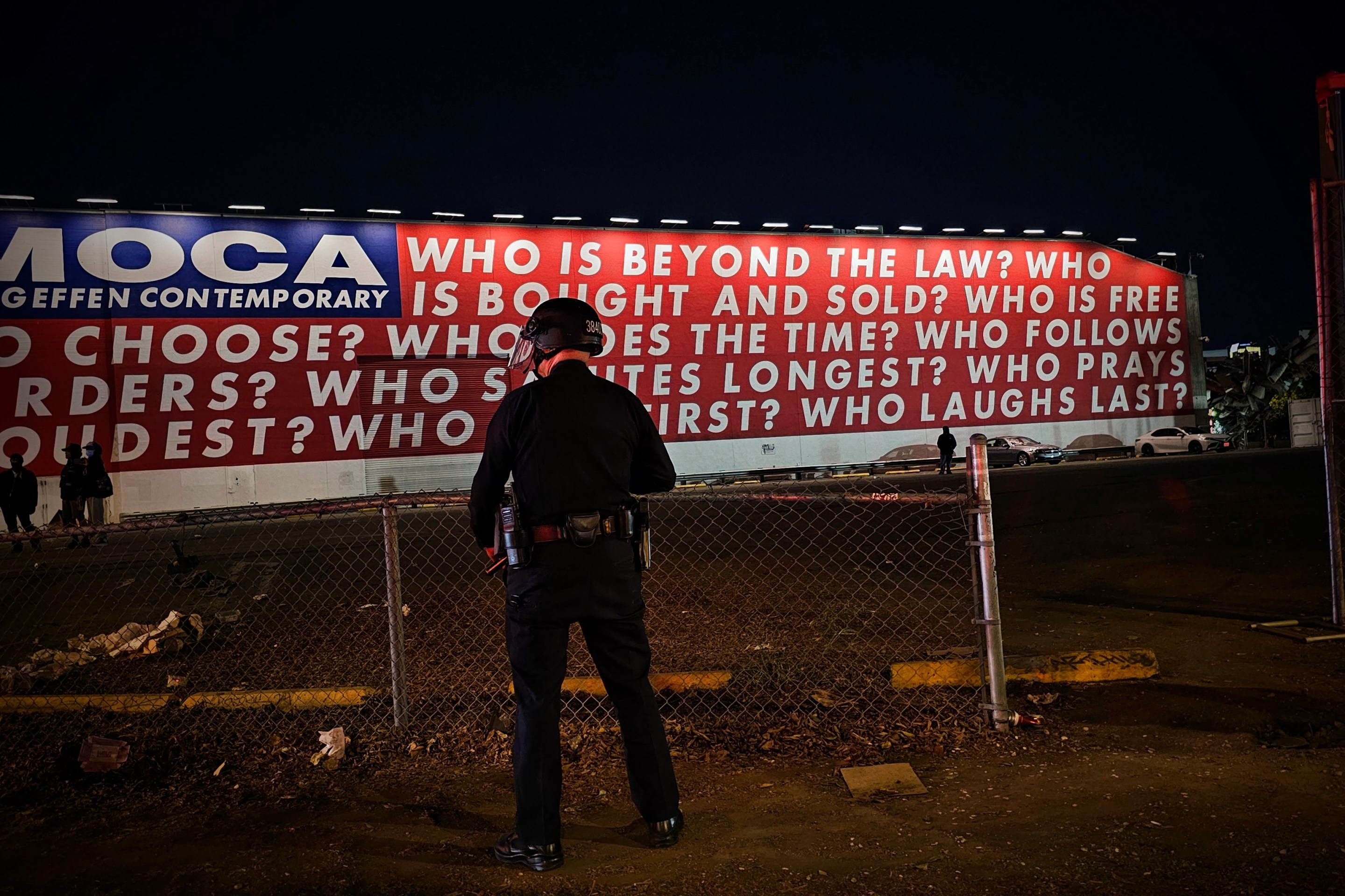 A Los Angeles police officer stands facing a red, white, and blue mural by the artist Barbara Kruger. The mural asks nine questions: “Who is beyond the law?”, “Who is bought and sold?”, “Who is free to choose?” “Who does the time?”, “Who follows orders?”, “Who Salutes longest?”, “Who prays loudest?”, “Who dies first?” and “Who laughs last?”.