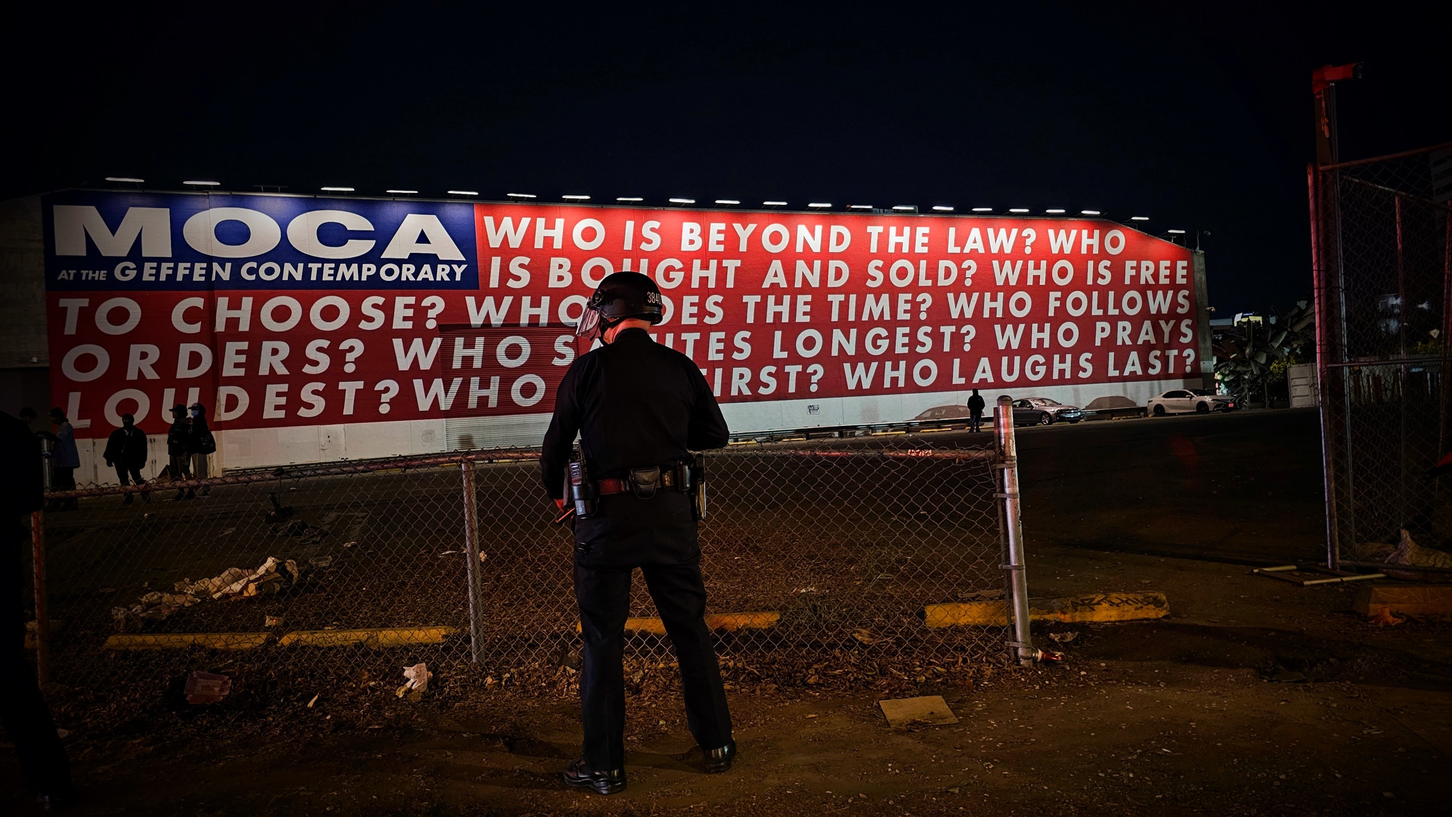 A Los Angeles police officer stands facing a red, white, and blue mural by the artist Barbara Kruger. The mural asks nine questions: “Who is beyond the law?”, “Who is bought and sold?”, “Who is free to choose?” “Who does the time?”, “Who follows orders?”, “Who Salutes longest?”, “Who prays loudest?”, “Who dies first?” and “Who laughs last?”.
