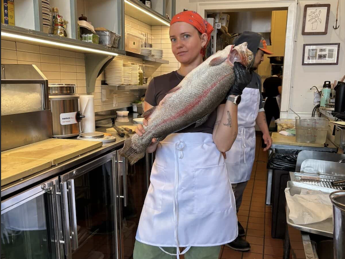 a woman holding a fish in a restaurant kitchen with other cooks in the back