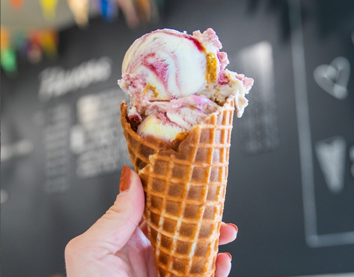 A hand with a red-painted thumbnail holds a waffle cone filled with scoops of a pink and white ice cream