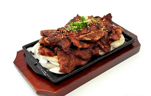 A plate of galbi topped with green onions and sesame seeds on a bed of onions, on top of a cast-iron plancha on a wooden board